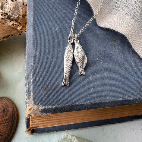 A silver fish pendant necklace on a delicate chain, displayed on top of a vintage blue book with a piece of burlap fabric and a seashell nearby.