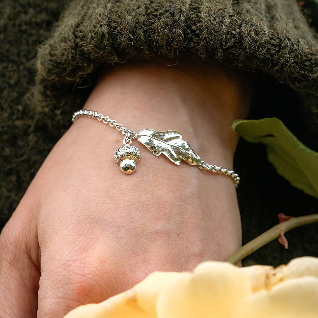 Hand wearing a silver bracelet with a yellow rose and green leaves in the background