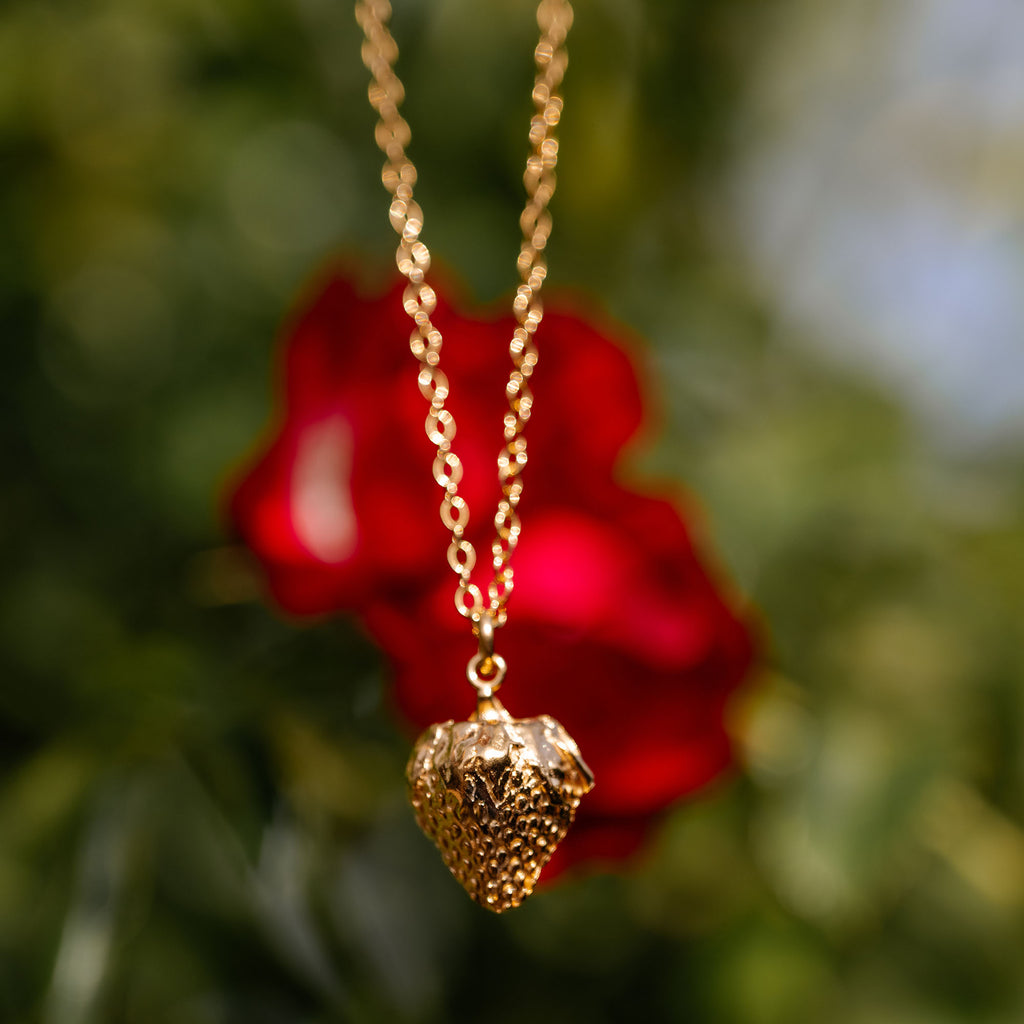 Gold necklace with a strawberry-shaped pendant against a blurred red flower background
