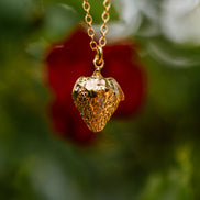 Gold strawberry-shaped pendant necklace against a blurred natural background