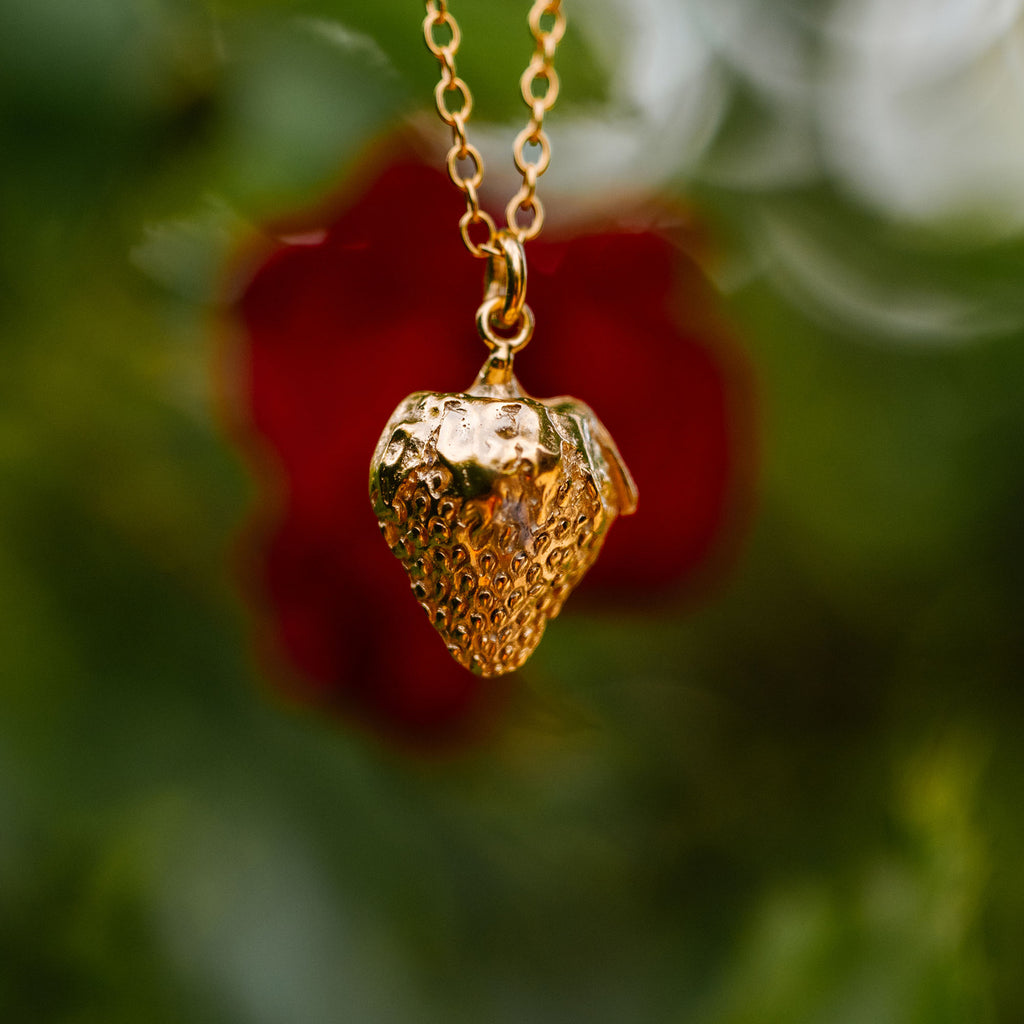 Gold strawberry-shaped pendant necklace against a blurred natural background