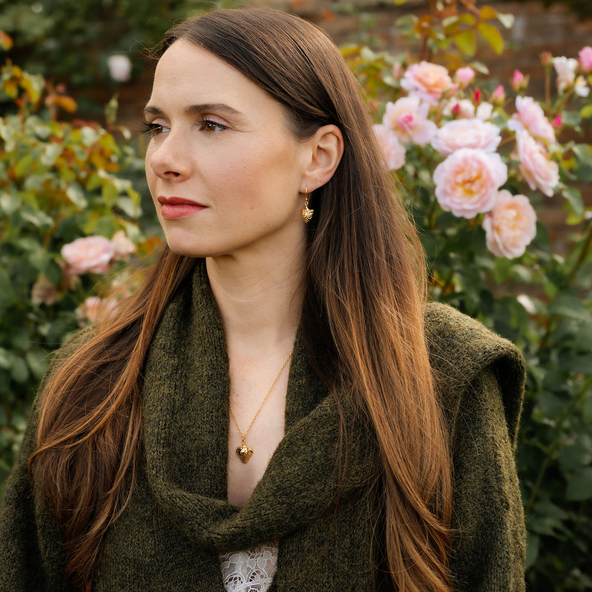 Woman with long brown hair wearing a green scarf and gold jewellery in a garden setting with pink flowers.
