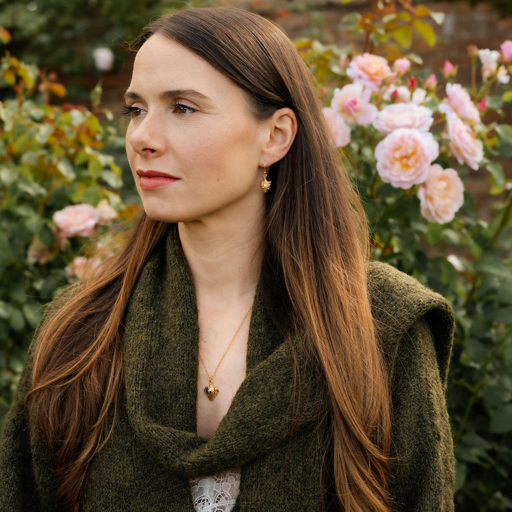 Woman with long brown hair wearing a green scarf and gold jewellery in a garden setting with pink flowers.